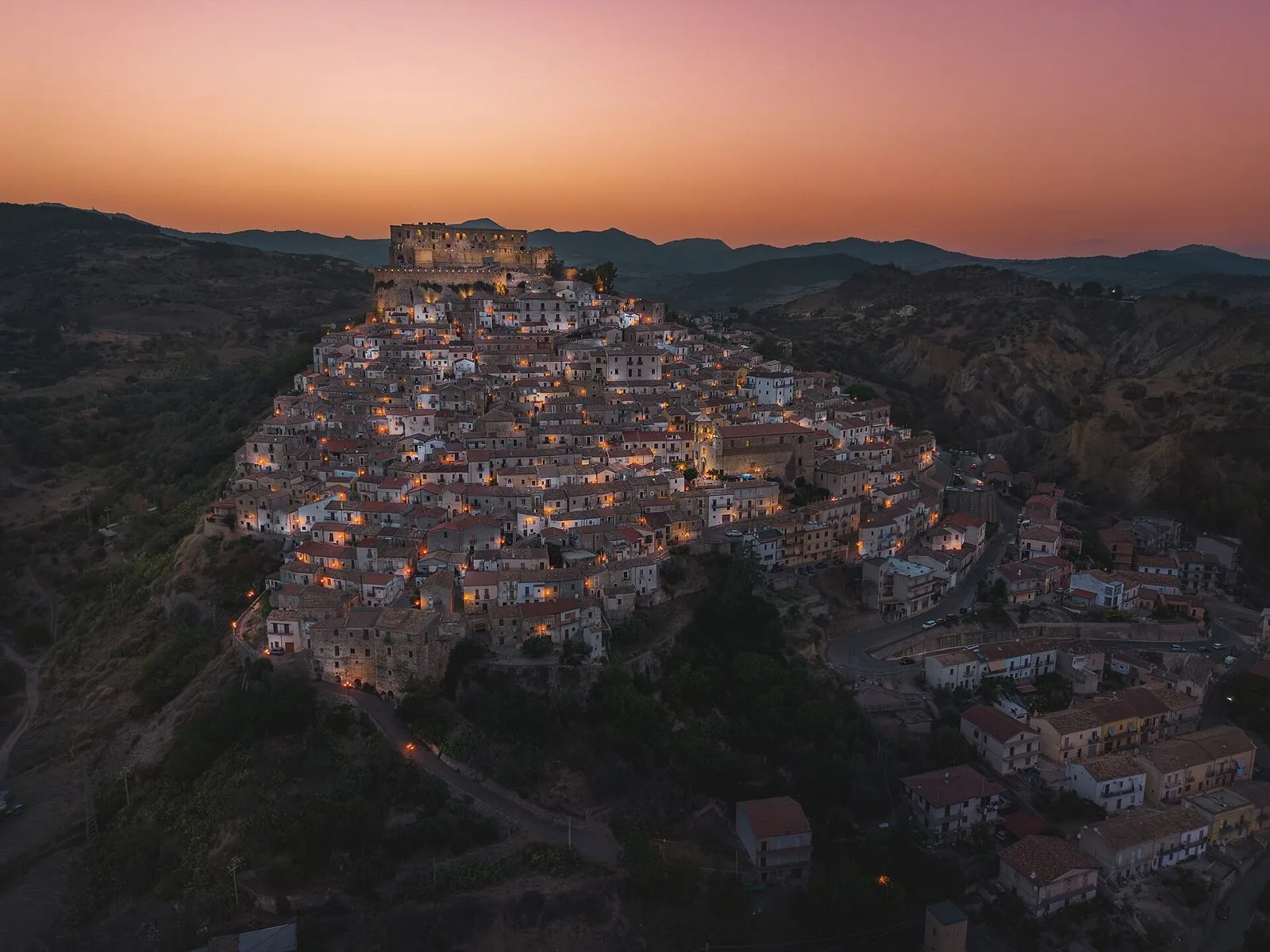 Veduta aerea al tramonto di Rocca Imperiale: le case del borgo illuminate salgono a gradinata sulla collina fino al Castello Svevo, con le montagne sullo sfondo
