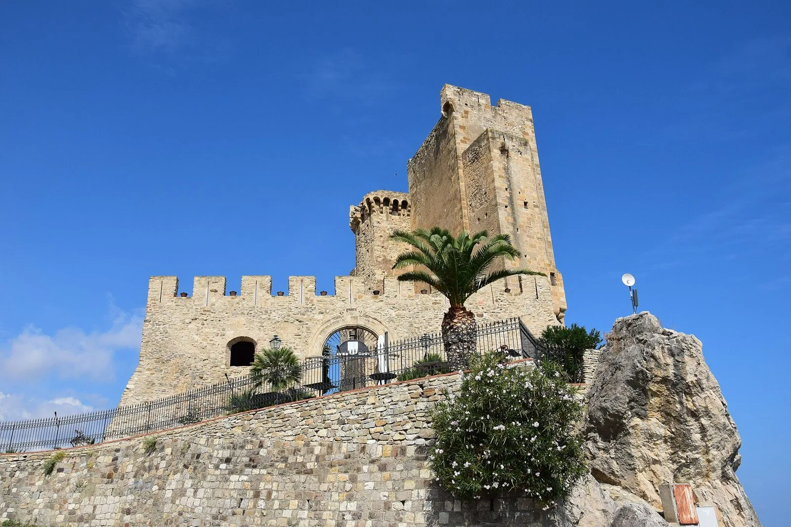 Il Castello Federiciano di Roseto Capo Spulico con la torre quadrata, le mura merlate, una palma e lo sperone di roccia sotto un cielo blu
