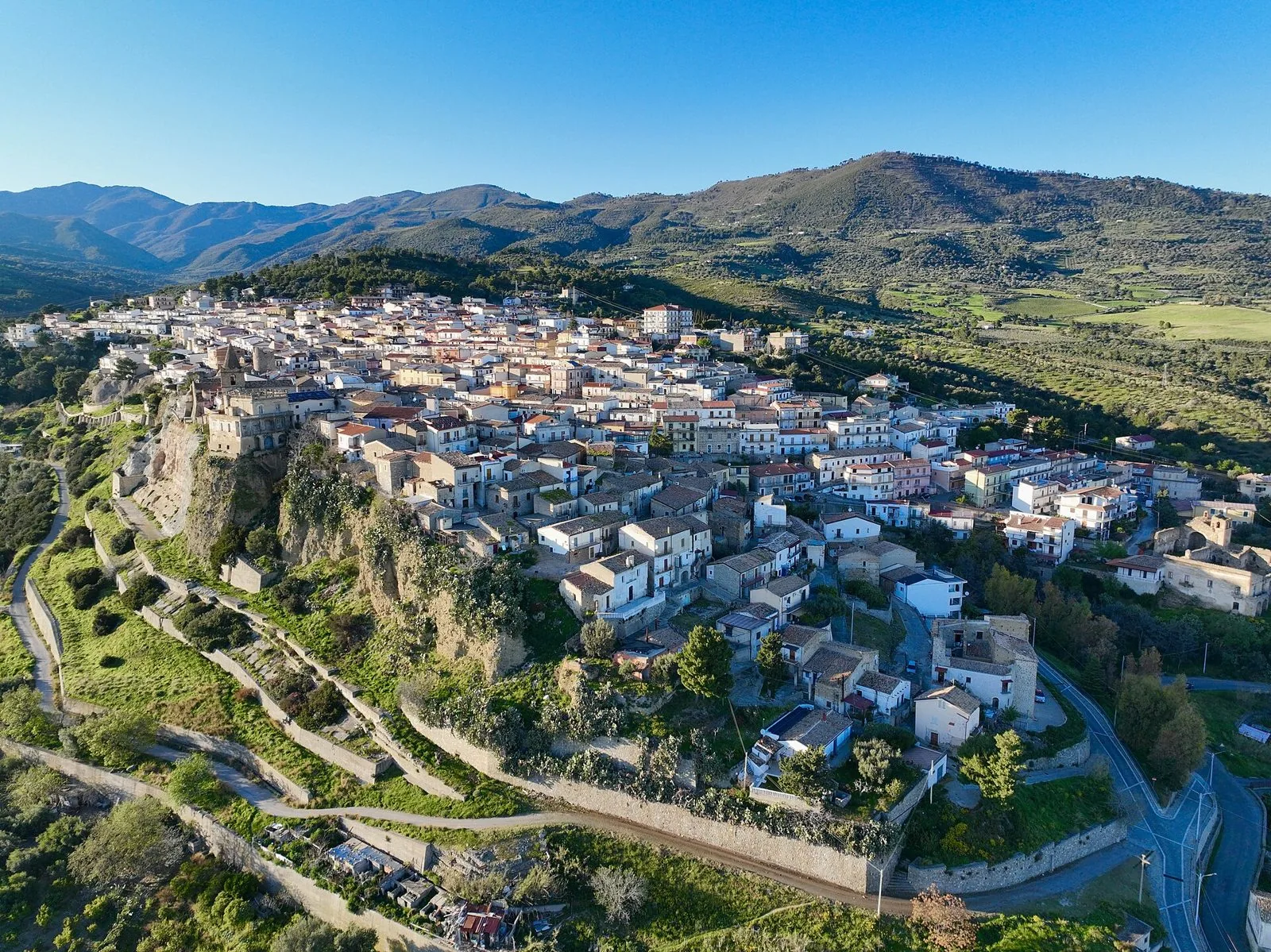 Veduta aerea del centro storico di Villapiana in una giornata di sole, con le case raccolte sul colle, i terrazzamenti verdi e le montagne sullo sfondo